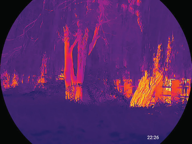 Circular thermal optic view of trees and landscape rendered in a purple, orange and yellow thermal colour palette with an on-screen time stamp
