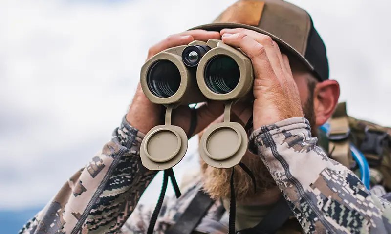 Hunter in camouflage holding binoculars while scanning open field terrain