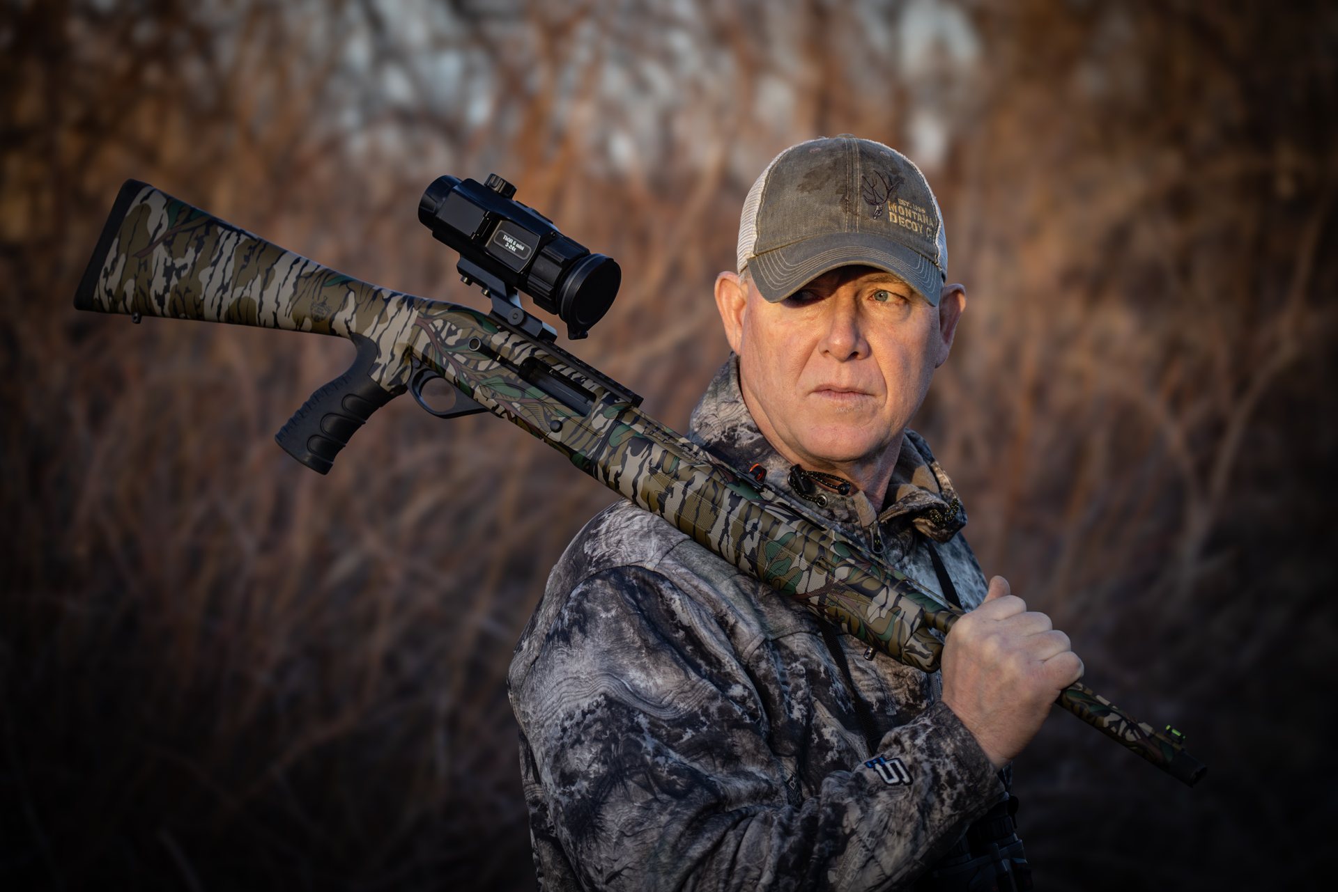 Hunter in camouflage resting a camouflage-patterned gun fitted with an ATN ThOR 6 Mini thermal scope over his shoulder in an open field